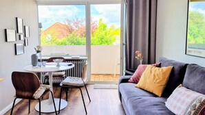 En moderne stue med en grå sofa, et spisebord med stole, en radiator og et stort vindue med udsigt over grøn vegetation og en bygning.