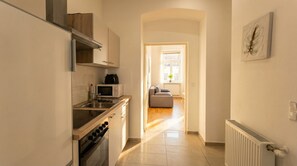Eine schmale Küche mit Mikrowelle, Herd und Spüle. Ein Durchgang führt zu einem Wohnbereich mit Sofa und Fenster.
