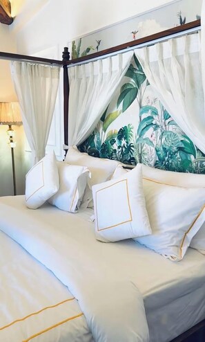 Uma cama com roupa de cama branca e detalhes dourados, um dossel com cortinas brancas leves e uma parede ao fundo com padrão de folhas tropicais.