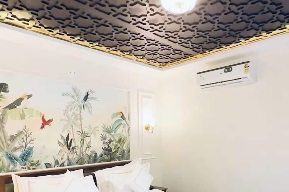 En pænt redt seng med hvide sengetæpper, en fastgjort aircondition og et loft med geometrisk mønster.