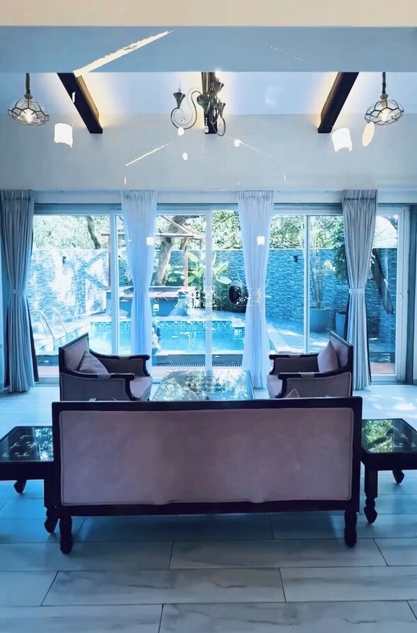 Uma sala de estar ampla com um sofá grande, uma mesa de centro de vidro e vista para uma piscina e áreas verdes.