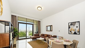 A modern living room with a sofa, dining table, and chairs, large windows with a city view, and a ceiling light. - Bloomfields Modern 1BR in Azizi Vista (Dubai)