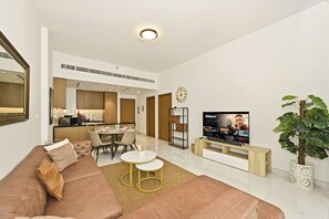 A modern living room with a sofa, TV, dining area, and kitchen in the background. - Bloomfields Modern 1BR in Azizi Vista (Dubai)