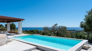 Una piscina rectangular con agua azul cristalina, rodeada por una terraza de baldosas blancas y una estructura de pérgola a la izquierda.