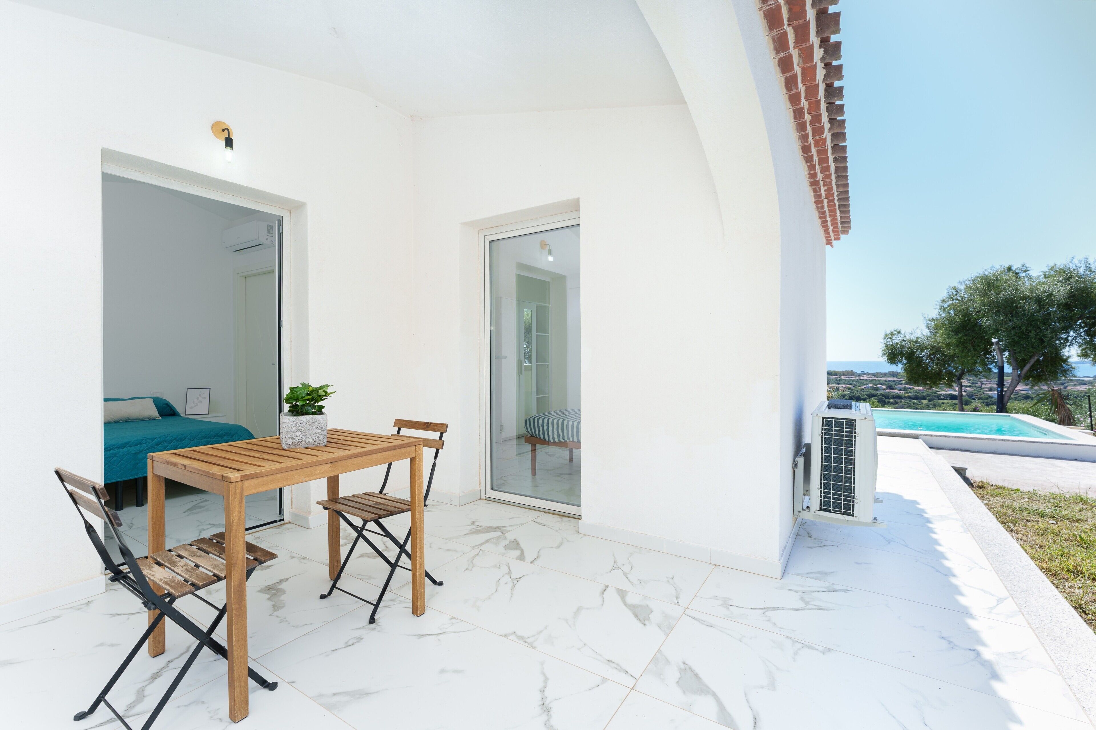 Um pátio com mesa e cadeiras de madeira, um pequeno vaso com planta e vista para uma piscina.