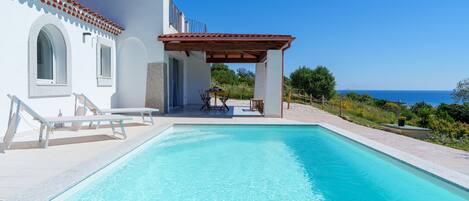 A poolside area with a clear view of the sea, a covered seating area, and a white building with a tiled roof.