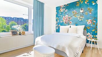 Un dormitori amb un llit gran, una finestra amb cortines, un radiador i una paret amb un mural floral.
