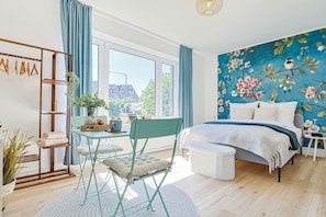 Een slaapkamer met een groot raam, een bed, een stoel, een tafel en een wand met een bloemmotief.