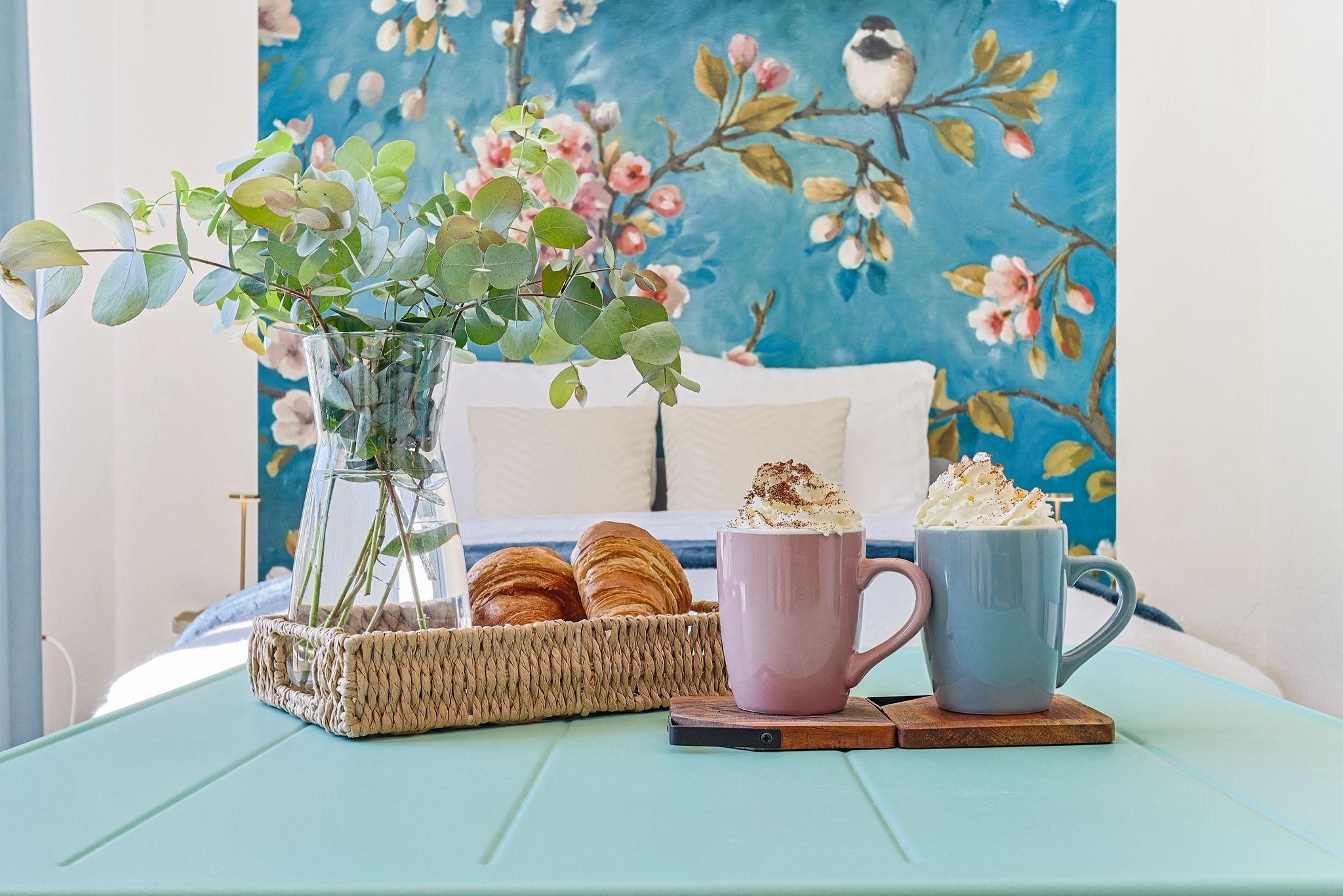 Una habitació amb un llit, un ram de fulles verdes, un cistell de croissants i dues tasses sobre una taula.