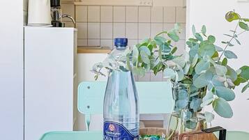 Een keukenhoek met een witte koelkast, een blauwe tafel, een waterfles, een vaas met groene bladeren en twee fotolijstjes aan de muur.