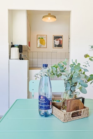 Een keukenhoek met een witte koelkast, een blauwe tafel, een waterfles, een vaas met groene bladeren en twee fotolijstjes aan de muur.