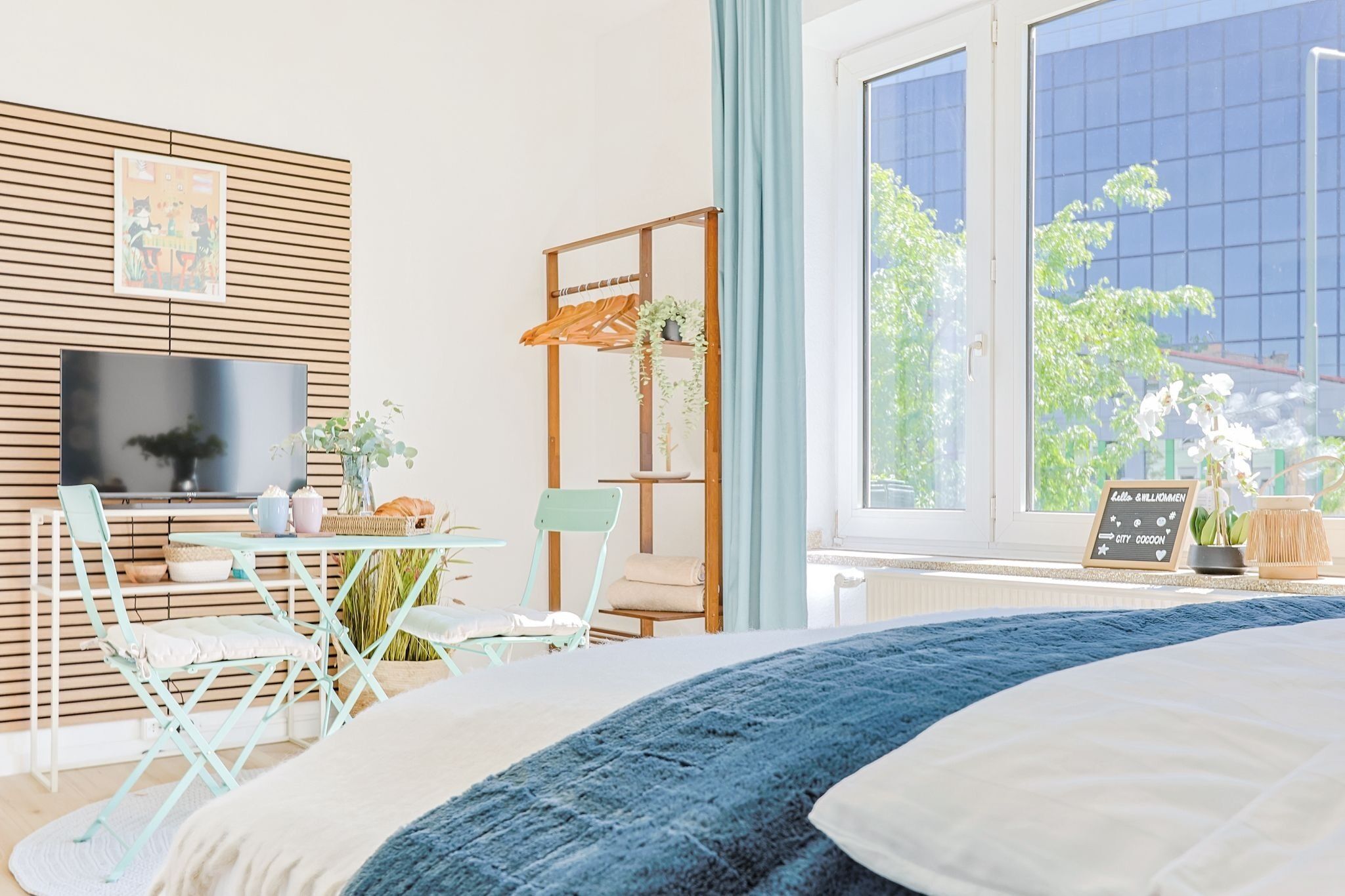 Ein Schlafzimmer mit einem großen Bett, einem Fernseher, einem Schreibtisch mit Stühlen, einem Fenster mit Blick auf Bäume und einem Regal mit Pflanzen.