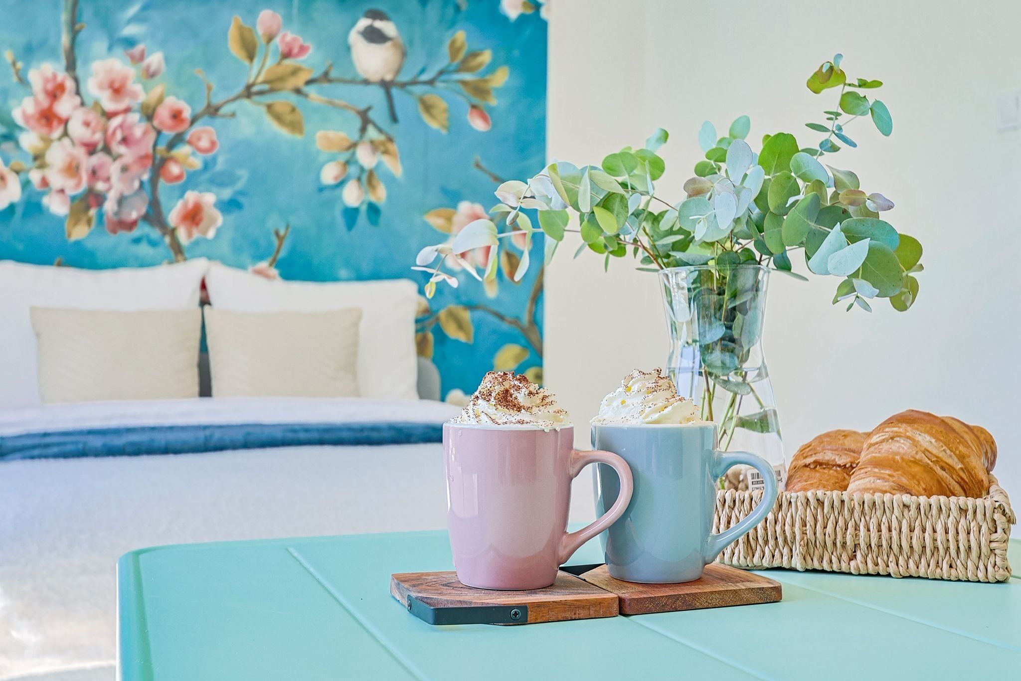 Ein Bett, ein Gemälde, eine Vase mit Pflanzen, zwei Tassen und ein Korb mit Brot auf einem Tisch.