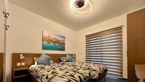 A bedroom with a bed, a nightstand, a wall-mounted lamp, a painting, and a window with blinds. - Luzzu Harbour Suites Modern 2BR in Marsaxlokk (Marsaxlokk)