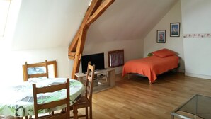 Ein Zimmer mit Holzbalkendecke, einem Bett mit orangefarbener Bettdecke, einem Fernseher auf einem Schrank, einem Tisch mit Stühlen und einem Fenster.
