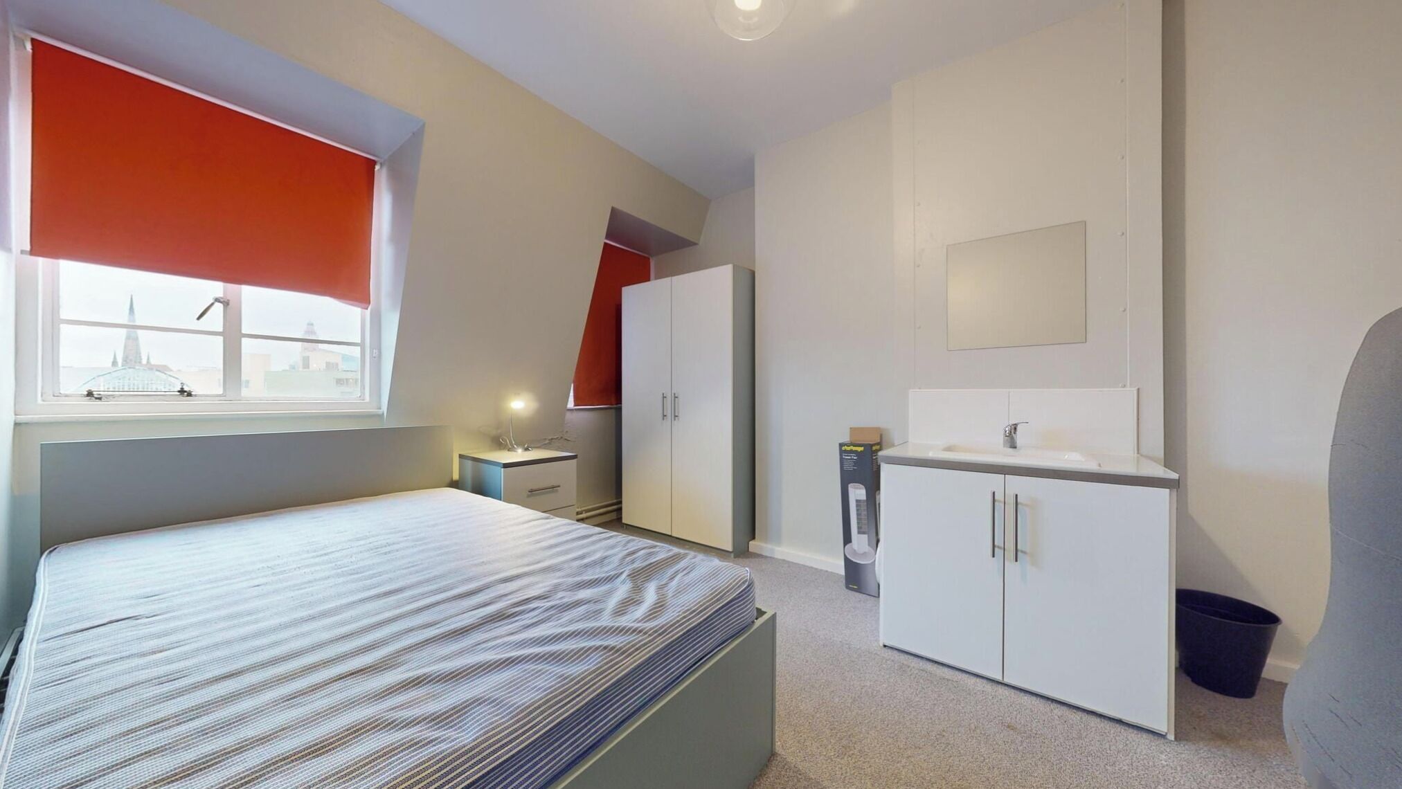 A bedroom with a bed, a window with a view, a wardrobe, a vanity with a mirror, and a trash bin.