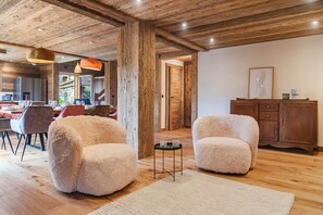 A spacious room with wooden beams, plush armchairs, a dining area, and a wooden cabinet.