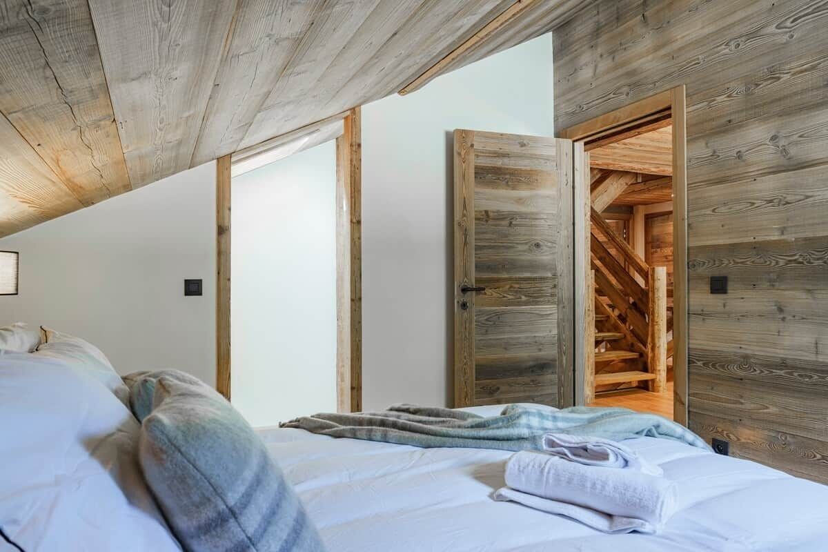 A bedroom with a wooden ceiling, a bed with white linens, and a wooden door leading to a staircase.