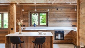 En stuga med träpanel på insidan, innehållande ett köksområde, ett matbord med stolar och ett fönster med utsikt över grönskan.