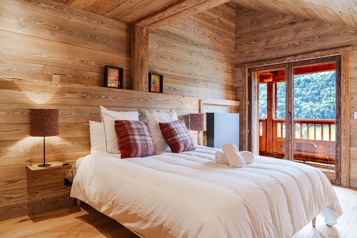 A wooden cabin-style bedroom with a large bed, two bedside lamps, and a window with a view of trees.