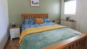 A bedroom with a wooden bed, green walls, a framed picture, a nightstand, a drawer, a window with a plant, and a potted plant on the nightstand. - Trillo Lodge (Colwyn Bay)