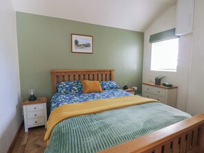 A bedroom with a wooden bed, green walls, a framed picture, a nightstand, a drawer, a window with a plant, and a potted plant on the nightstand. - Trillo Lodge (Colwyn Bay)