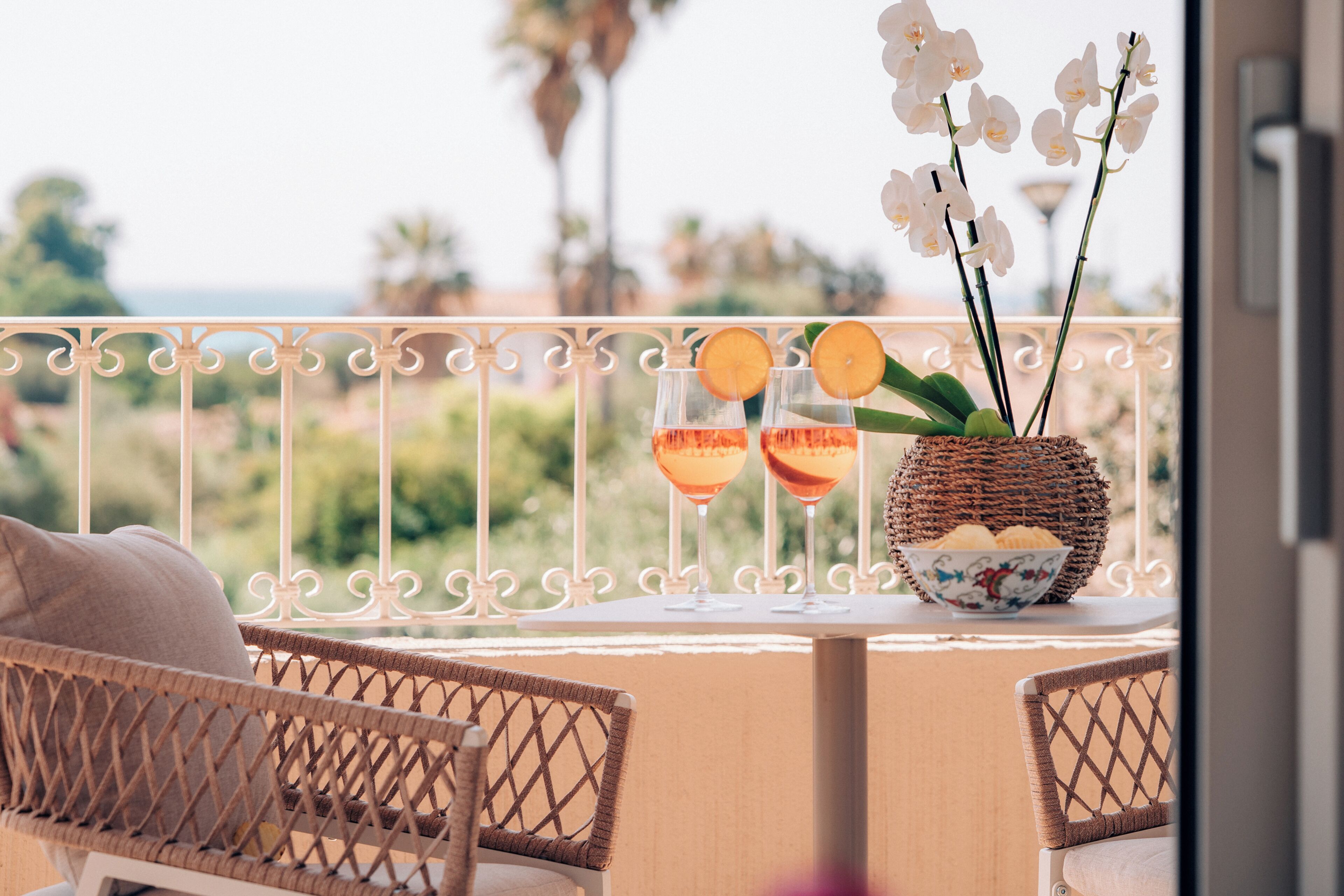 Ein Balkon mit Korbstühlen, einem Tisch mit zwei Gläsern rotem Getränk, einem Korb voller Blumen und einer Schüssel mit Snacks.