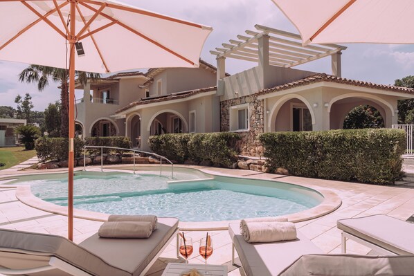 A swimming pool with lounge chairs and umbrellas, surrounded by a landscaped garden and a multi-story building with arches. - Lentischi Hotel - Adults Only (Simius)