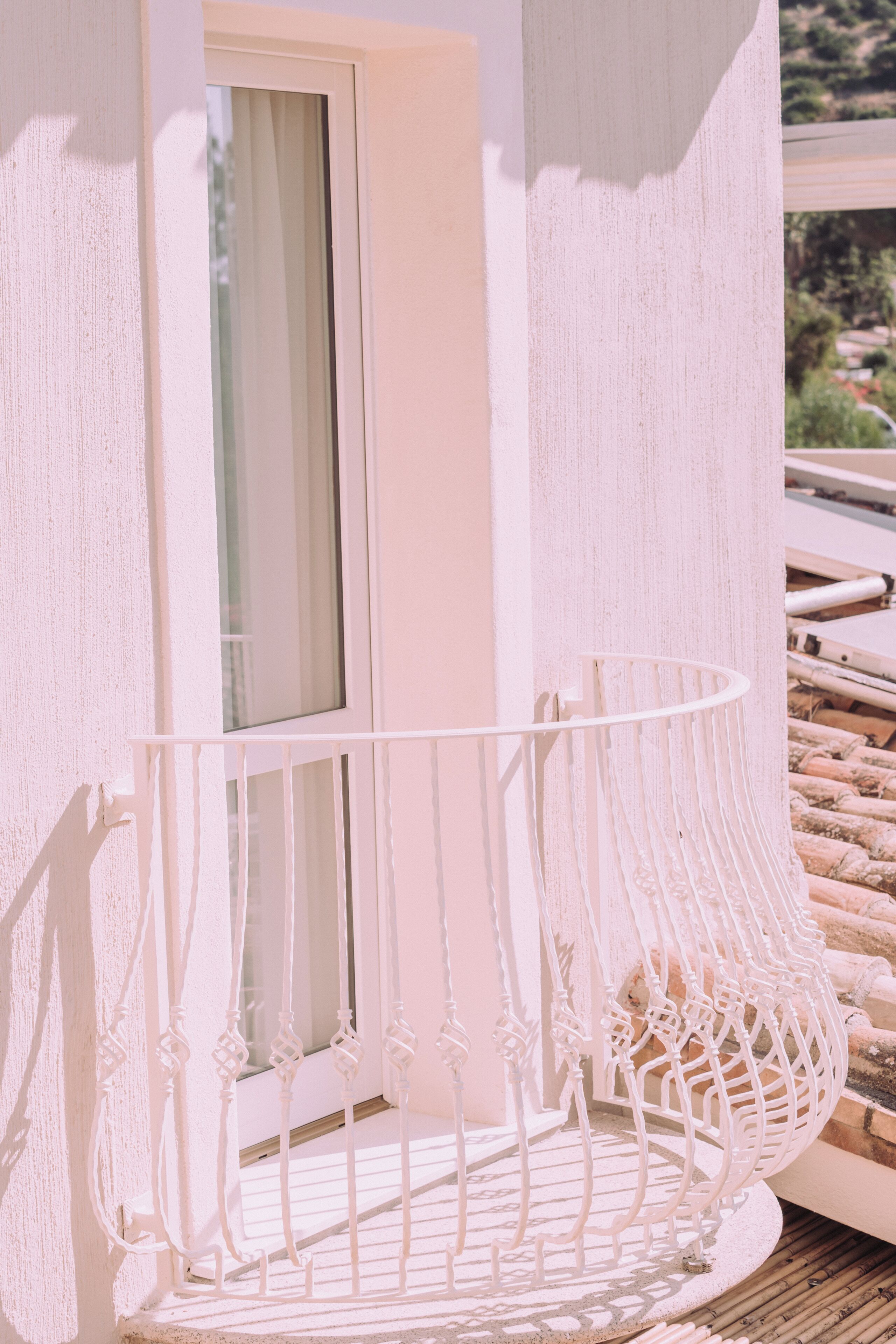 A white balcony with a wrought iron railing, a window, and a view of a hillside.