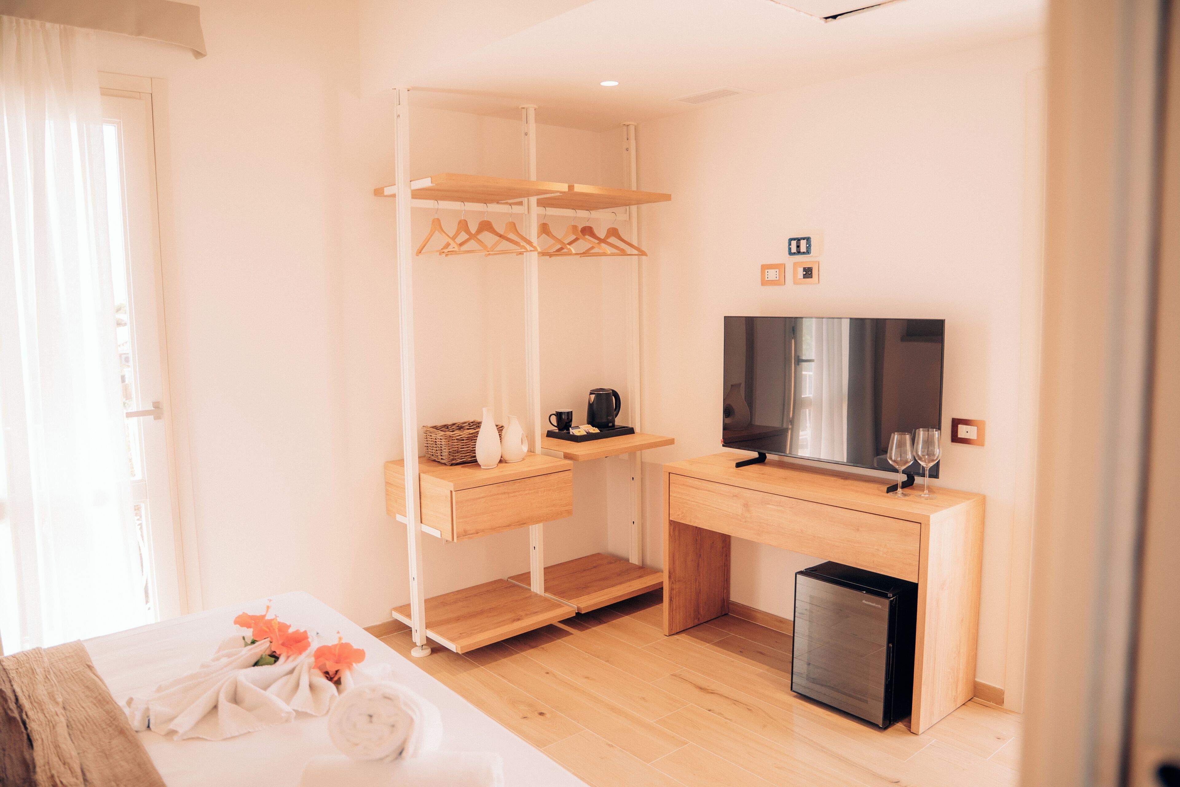 Quarto de hotel com cama, televisão, mini-geladeira, escrivaninha de madeira e guarda-roupa com cabides.