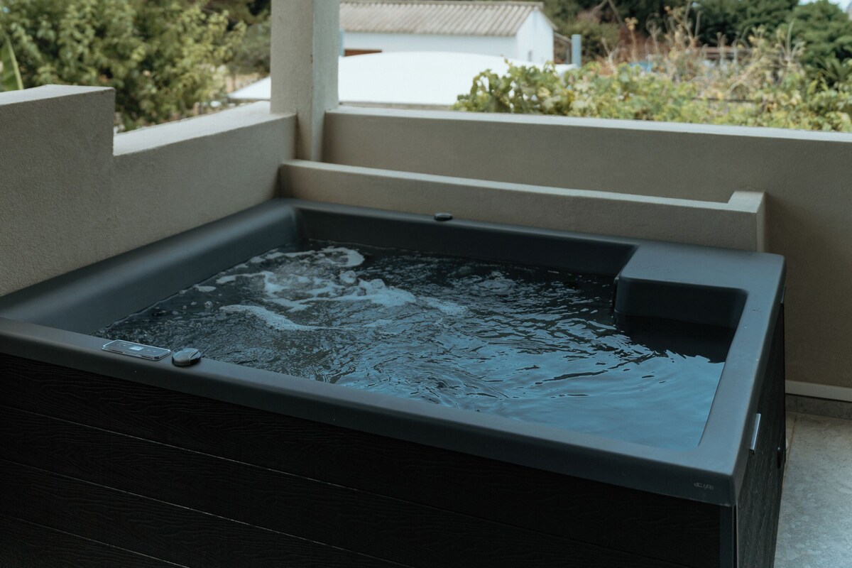 Un jacuzzi bajo una zona techada con vistas a una casa y árboles al fondo.