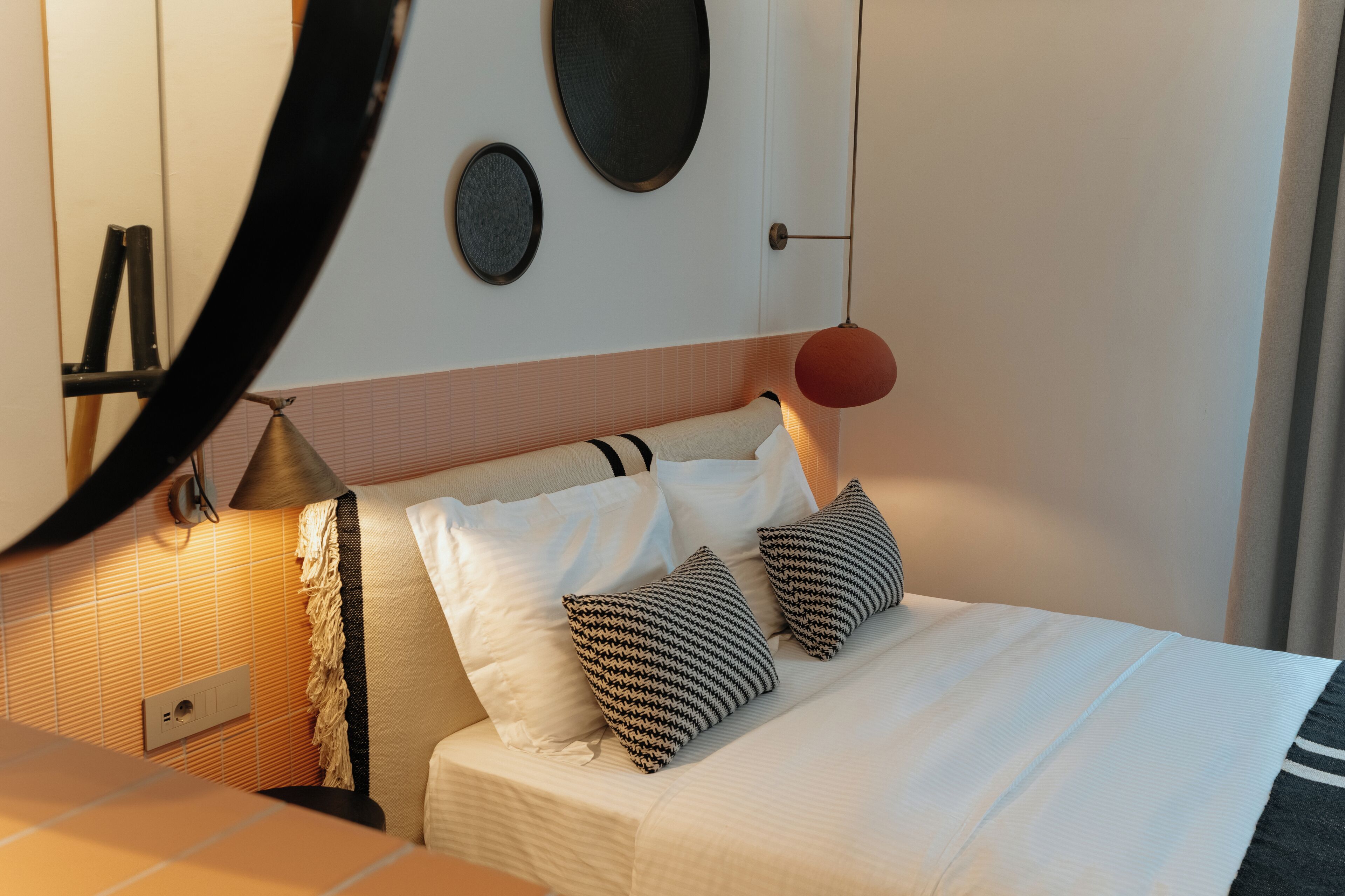 Un lit bien fait, avec une literie blanche et deux coussins à motifs, une lampe de chevet et un miroir rond fixé au mur.