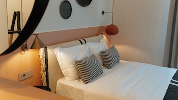 Un lit bien fait, avec une literie blanche et deux coussins à motifs, une lampe de chevet et un miroir rond fixé au mur.