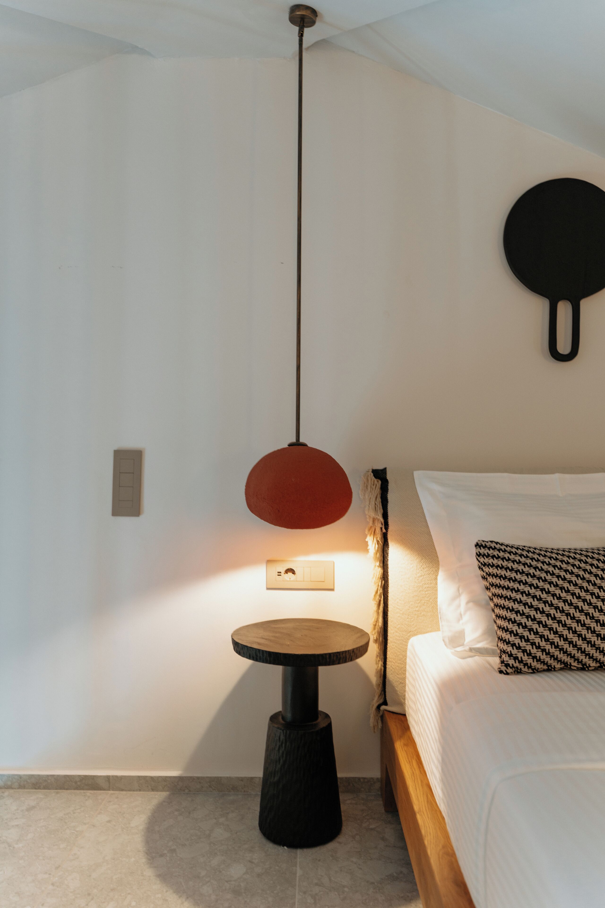 Un dormitorio con una cama, una mesita redonda, una lámpara colgante y un objeto sujeto a la pared.