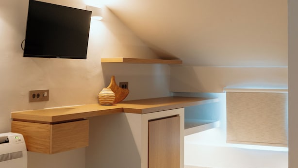Ein modernes Schlafzimmer mit schrägem Dach, einem an der Wand montierten Flachbildfernseher, einem Nachttisch aus Holz und einer Nachttischlampe sowie einem schwarzen Teppich.