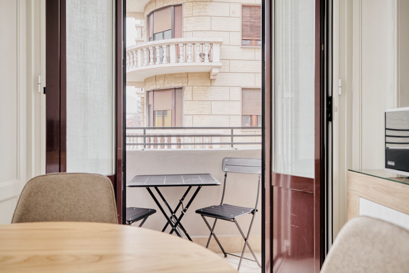 A room with a wooden table, two chairs, a glass partition, and a view of a building with balconies.