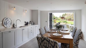 A modern kitchen with white cabinets, a dining table set for four, and a view of a garden through a large window. - Rose Cottage (Wadebridge)