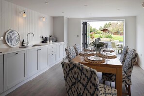 Una cocina moderna con armarios blancos, una mesa de comedor lista para cuatro personas y vistas a un jardín a través de un gran ventanal.