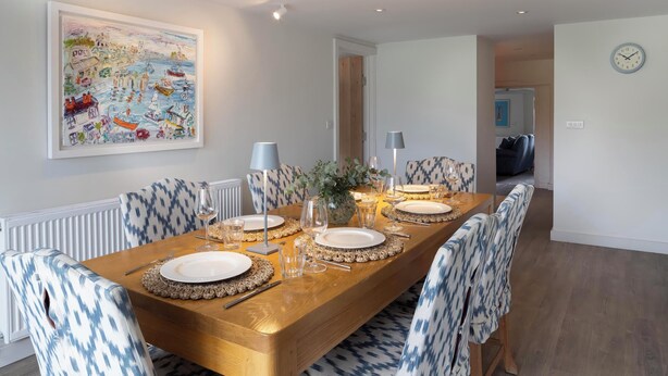 A dining room with a long wooden table set for four, patterned chairs, a framed painting, and a clock on the wall. - Rose Cottage (Wadebridge)