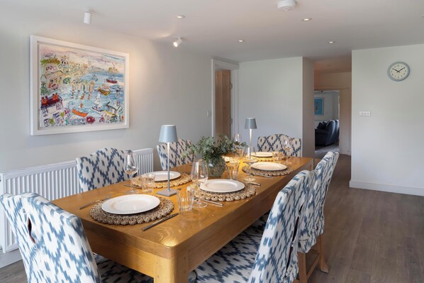 A dining room with a long wooden table set for four, patterned chairs, a framed painting, and a clock on the wall.