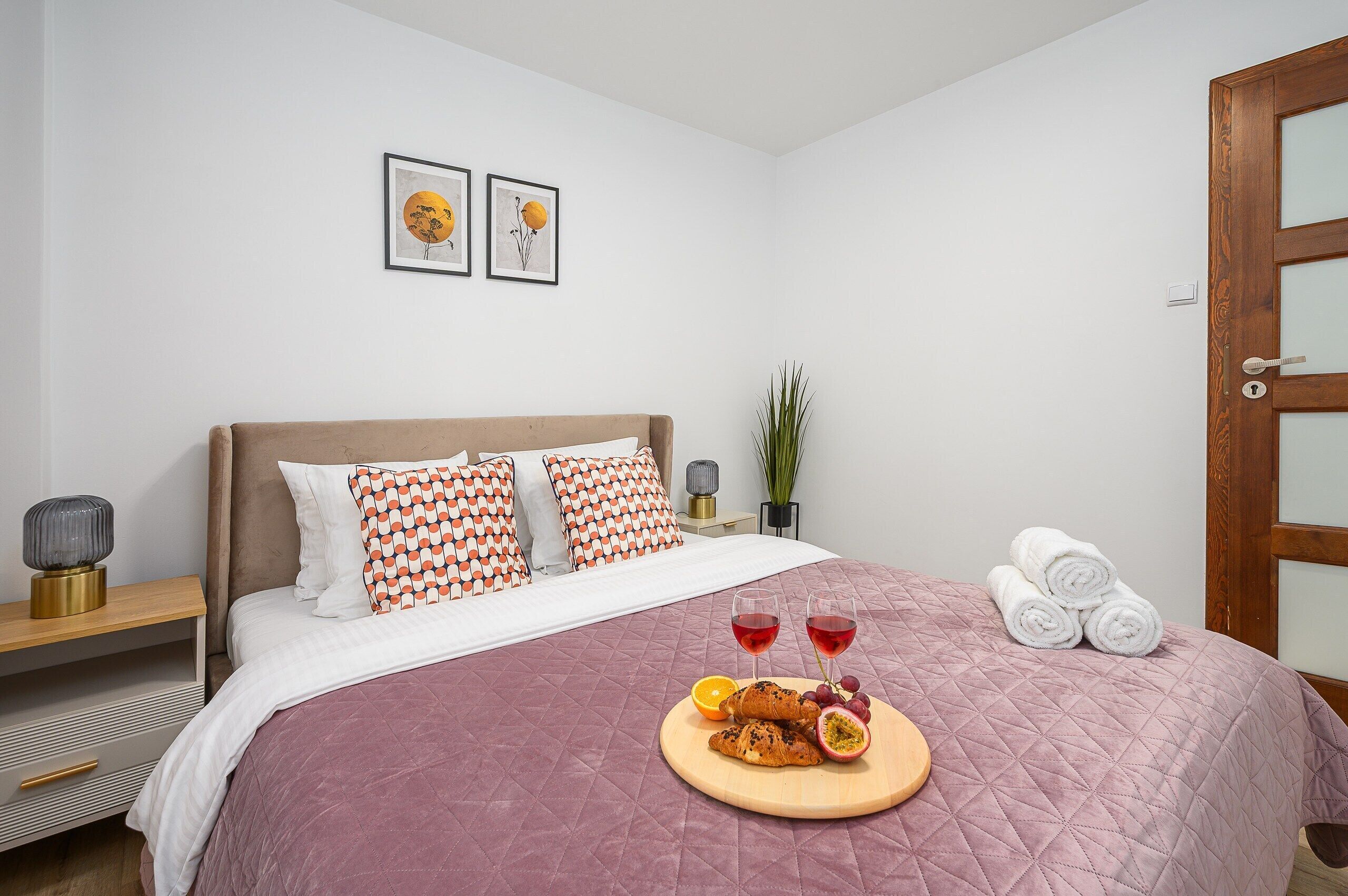 Una cama bien hecha con colcha rosa, una mesita de noche con lámpara y una bandeja con comida y bebidas.