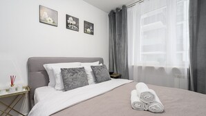 En pænt redt seng med sammenrullet håndklæder, et natbord med lampe, og et vindue med gardiner.