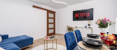 Uma sala de estar moderna com um sofá seccional, uma mesa de jantar posta para dois, uma TV de tela plana exibindo 'NETFLIX' e uma porta de madeira.