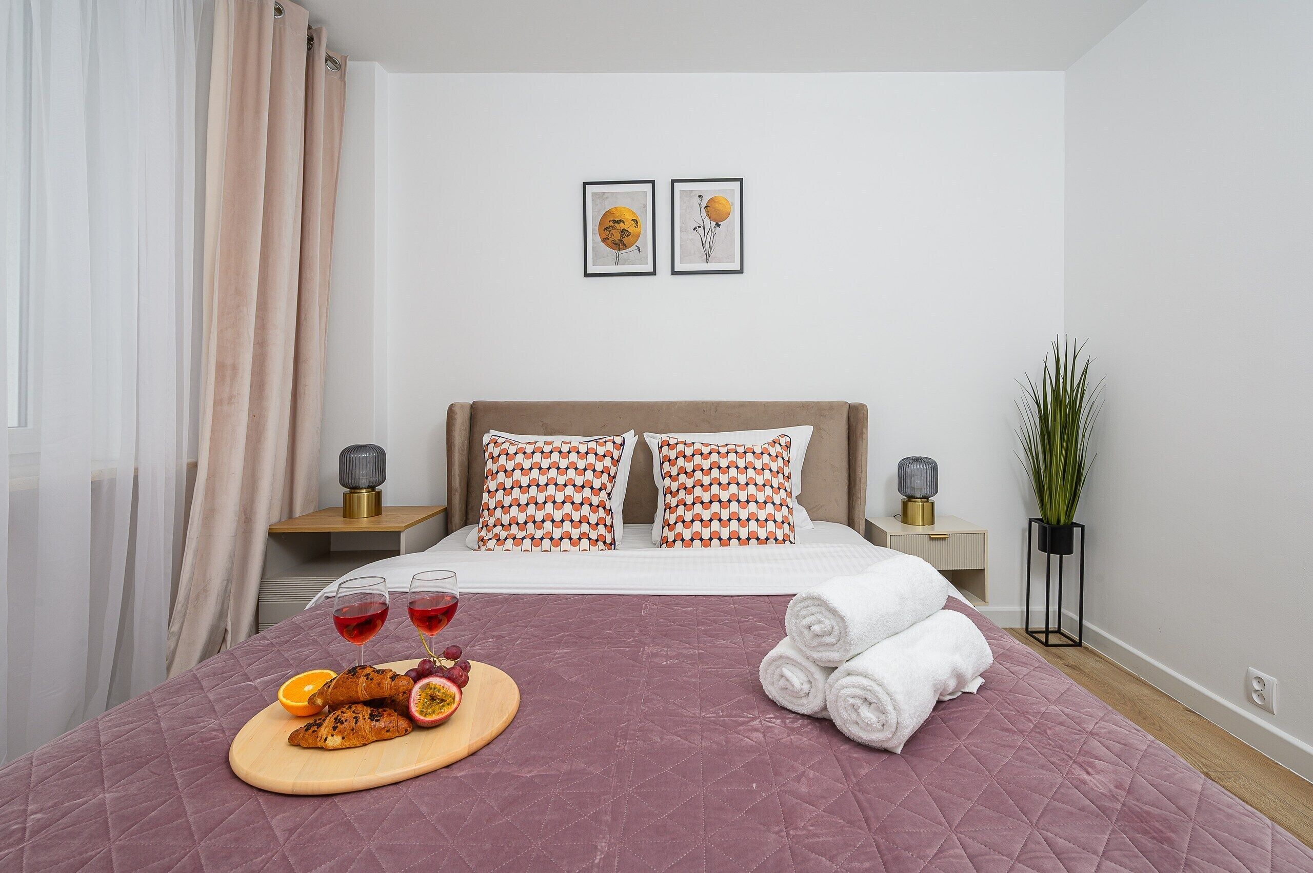 Ein ordentlich eingerichtetes Hotelzimmer mit einem Bett, Kissen, einem Nachttisch mit Lampe, einer kleinen Topfpflanze und einem Frühstückswagen mit Croissants und Obst.