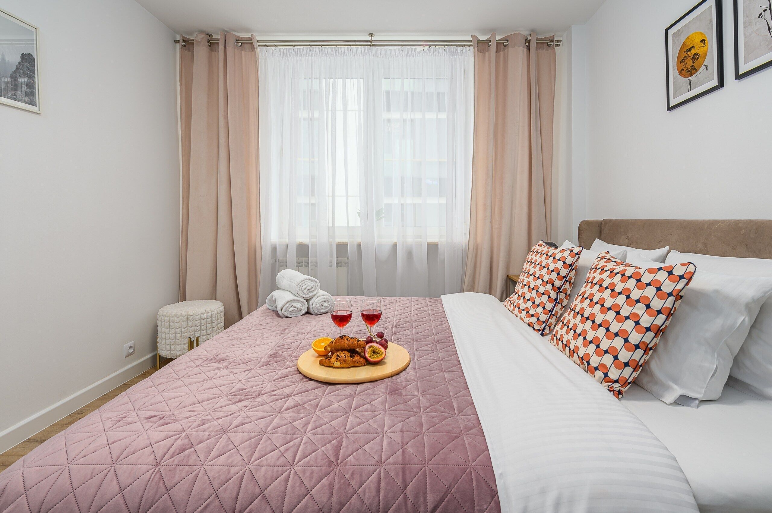 Ein ordentlich gemachtes Bett mit einer rosa Steudecke, ein Holztablett mit Snacks und Getränken und Blick auf die Stadt durch leichte Vorhänge.