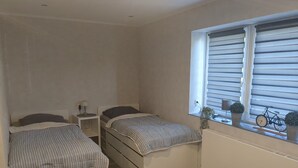 Une chambre avec deux lits, une fenêtre avec des stores, un radiateur et une petite chaise.