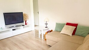 Un salón con televisión de pantalla plana, una mesa de centro blanca, un sofá beige con cojines y una planta pequeña sobre un mueble blanco.