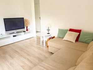 Ein Wohnzimmer mit Flachbildfernseher, einem weißen Couchtisch, einem beige farbenen Sofa mit Kissen und einer kleinen Pflanze auf einer weißen Konsole.