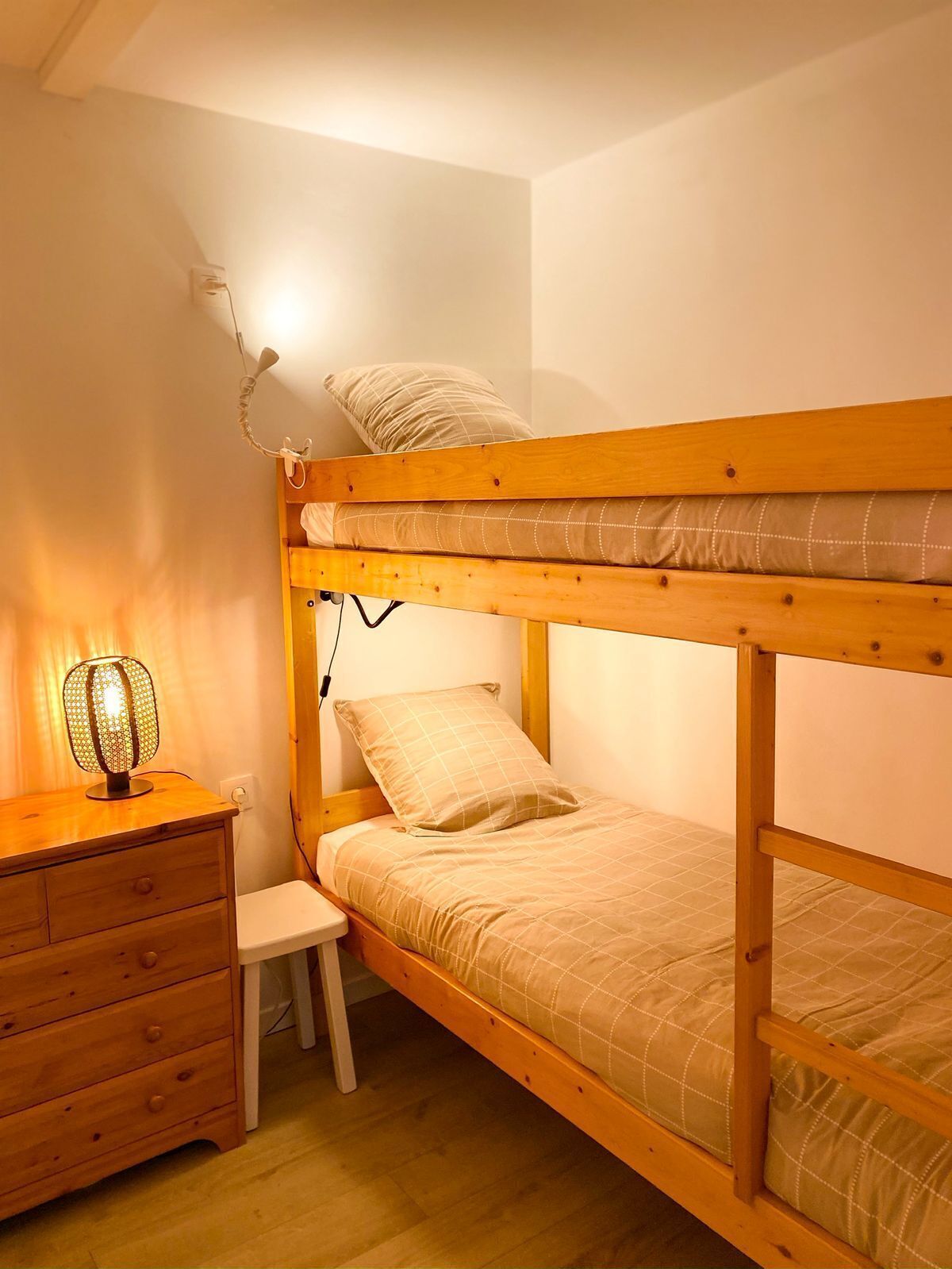 A bunk bed with a wooden frame, a bedside table with drawers, a small chair, and a wall-mounted lamp.