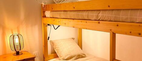 A bunk bed with a wooden frame, a bedside table with drawers, a small chair, and a wall-mounted lamp.
